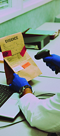 laboratory technician, wearing gloves, holding sealed evidence bag and barcode scanner, scanning item of evidence into inventory on laptop computer.