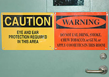 Signs on door to test firing range. Sign one reads, 'CAUTION: Eye and ear protection required in this area.' Sign two reads, 'WARNING: Do not eat, drink, smoke, chew tobacco, or gum, or apply cosmetics in this room.'
